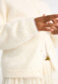 Cardigan morbido di colore crema, con una consistenza soffice, dotato di chiusura a bottoni e polsini a coste, indossato sopra una gonna con bordi in pizzo.