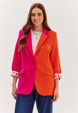 Blazer s farebnými blokmi, ktorý obsahuje ružové a oranžové sekcie, s manžetou posiatou bodkami. Výrazný srdcový detail na náprsnom vrecku.