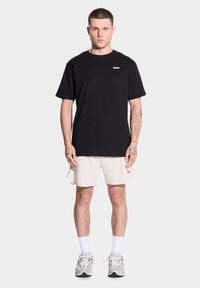 Młody mężczyzna o krótkich włosach stoi przodem, ubrany w czarną koszulkę i kremowe szorty, w połączeniu z białymi skarpetkami i sportowymi butami.