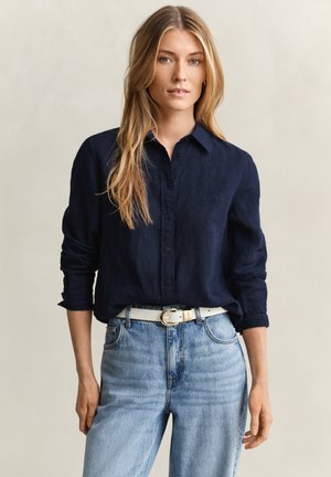 Blonde Frau mit langem Haar, die ein marineblaues, knöpfbares Hemd trägt, das in hellblaue Jeans mit weißem Gürtel gesteckt ist, steht vor einem einfarbigen Hintergrund.