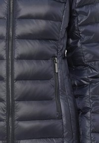 Ναυτική επένδυση μπουφάν με πλισέ σχέδιο, διαθέτοντας μπροστινό φερμουάρ και πλαϊκή τσέπη με φερμουάρ. Το υλικό έχει γυαλιστερή επιφάνεια και λεία υφή.