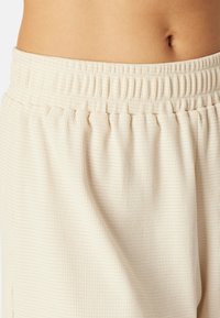 Nahaufnahme von der Taille einer Person, die cremefarbene, elastische Hosen mit Textur trägt, wobei Haut und Bauchnabel oberhalb der Hosen sichtbar sind.