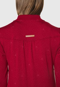 Pull rouge avec un motif texturé et un petit logo au niveau du col. Il présente un col côtelé et un pli piqué à l'arrière.