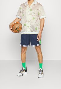 Hedvábná košile s krátkým rukávem se vzorem palem v světle béžové barvě, kombinovaná s námořnickými kraťasy s zelenými akcenty a bílými pruhy. Zelené ponožky a sporty boty.