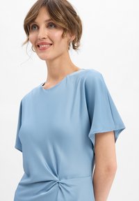 Femme aux cheveux courts et ondulés, portant un haut léger bleu clair à manches courtes avec un détail de nœud tordu à la taille, souriante et regardant légèrement vers le haut.