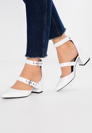 Fødder iført hvide sko med blokhæle og dobbelte spænder, kombineret med mørkeblå, korte jeans med frynser ved kanterne.