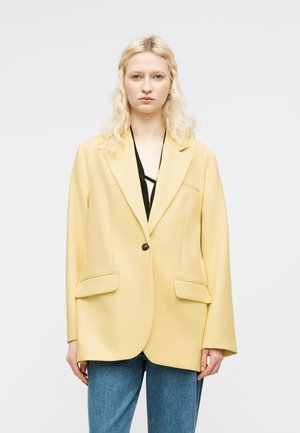 Γυναίκα με ξανθά μαλλιά, φοράει ένα χαλαρό απαλό κίτρινο σακάκι πάνω από μαύρη μπλούζα και μπλε τζιν, στέκεται μπροστά σε απλό ανοιχτόχρωμο φόντο.