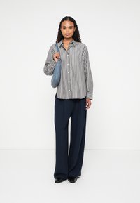 Camicia a righe grigia e bianca, abbinata a pantaloni larghi blu navy. Tiene in mano una borsa azzurro chiaro. Tessuto liscio, vestibilità rilassata.