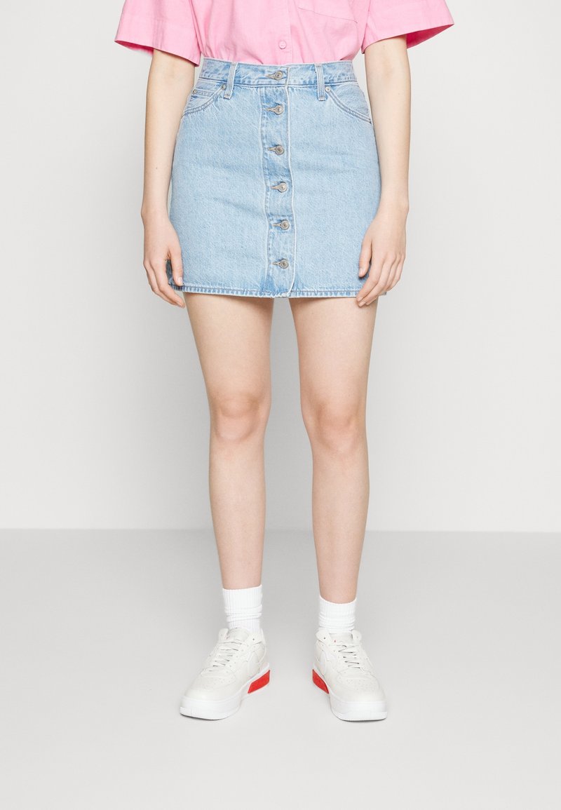 Personne portant une jupe en denim bleu clair à boutonnière devant, des chaussettes blanches, des baskets blanches avec des touches rouges et un t-shirt rose à manches courtes.