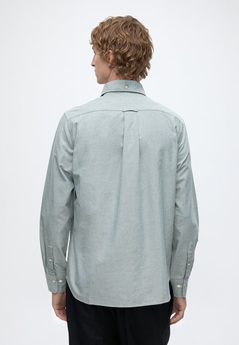 Hombre con cabello rizado que lleva una camisa de botones de manga larga color gris claro visto desde atrás con un fondo blanco liso.