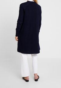 Persona vestida con un largo cárdigan azul marino, pantalones blancos y zapatos negros sin cordones, de pie de espaldas en un fondo gris claro liso.