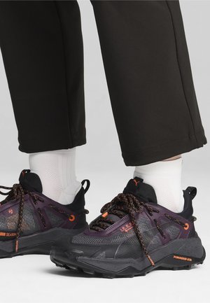Czarno-fioletowe sneakersy mają teksturowany wzór, siateczkowe wstawki oraz pomarańczowe akcenty. Sznurówki są pomarańczowe z odblaskowym detalem.
