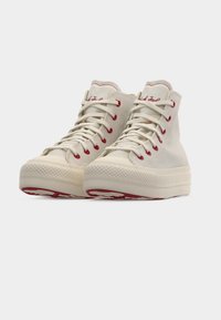 Κρεμ καμβαδένια αθλητικά παπούτσια υψηλού μπροστινού μέρους με παχιά λαστιχένια σόλα, κόκκινα τρυπήματα και λευκά κορδόνια, σε λευκό φόντο.