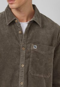 Bruine corduroy blouse met knoopsluiting aan de voorkant, borstzak en een klein logopatch. Gestructureerde stof met verticale ribbels.