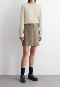 Frau in einer cremefarbenen Langarmbluse, brauem knielangen Rock, schwarzen Socken und schwarzen Plateauslippern, die auf einem grauen Boden steht.