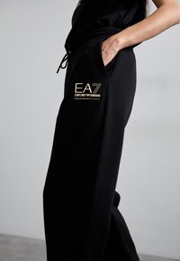 Zwarte joggers met een trekkoord in de taille hebben een glanzend gouden EA7 Emporio Armani-logo op de linkerzak, een gladde stoftextuur en een casual ontwerp.