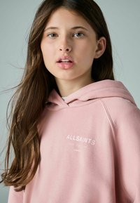 Růžová mikina s kapucí na šňůrku, s bílým logem "ALLSAINTS". Měkký materiál, volný střih, mírně viditelná textura.