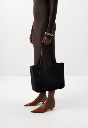 Person in a brown ribbed dress holding a large black tote bag, wearing a chunky gold bracelet and brown pointed low-heeled shoes.