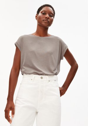 Vrouw staand met hand op heup, draagt een los grijs kort mouwtopje en witte broek met hoge taille tegen een effen achtergrond.