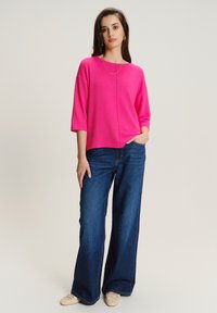 Kobieta stoi z jedną ręką w kieszeni, ubrana w jasnoróżowy prążkowany top, szerokie niebieskie dżinsy, beżowe baleriny i złotą biżuterię.