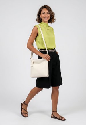 Lachende vrouw die loopt, draagt een limegroene mouwloze top, zwarte shorts, bruine sandalen en draagt een witte schoudertas.