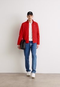 Rode gestikte jas, witte polo, blauwe jeans en witte sneakers. Een zwarte pet en een zwarte schoudertas maken de informele outfit compleet.