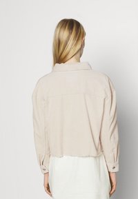 Žena s dlouhými blond vlasy otočená zády, oblečená v světle béžové zkrácené manšestrové bundě přes bílé šaty na jednolitém pozadí.