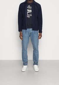 Chaqueta texturizada azul marino sobre camiseta gráfica negra, combinada con jeans azul claro y zapatillas blancas. Ajuste casual y relajado en general.