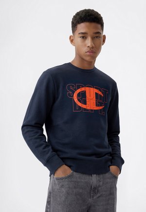 Giovane uomo con capelli corti e ricci che indossa una felpa blu navy con testo arancione "SPORT DEPT." e jeans grigi con le mani in tasca.