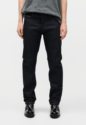 Άνδρας που φοράει μαύρο τζιν κανονικής γραμμής, μαύρα λαμπερά μπότες με φερμουάρ μπροστά και γκρι φθαρμένο t-shirt, στέκεται μπροστά από ένα απλό φόντο.