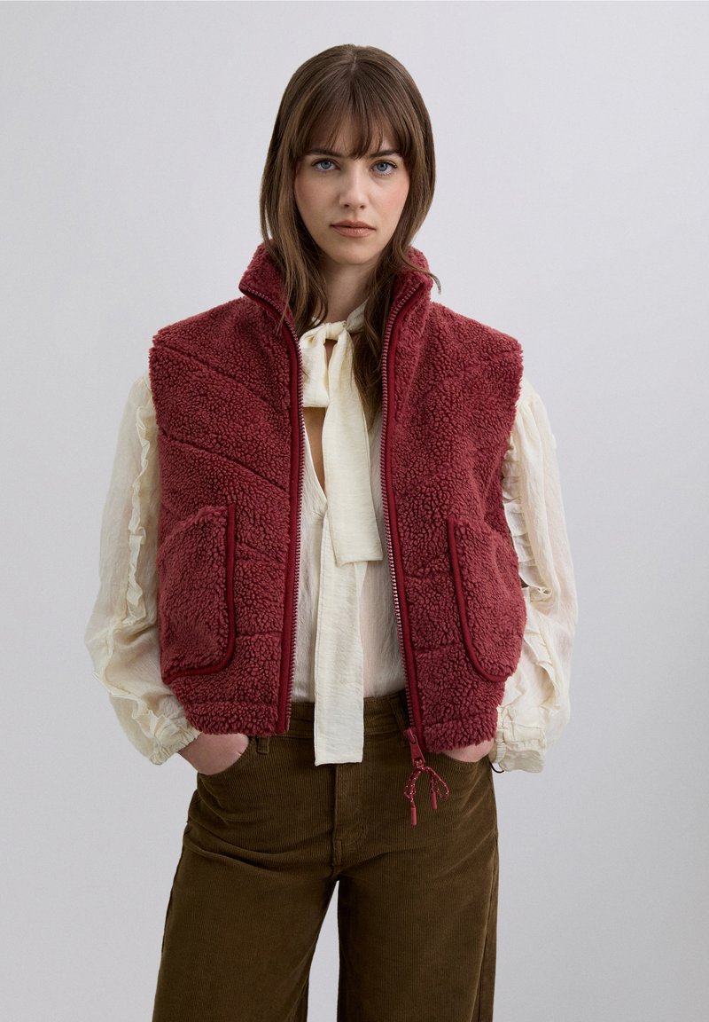 Chaleco de forro polar burdeos con cuello alto, dos bolsillos frontales y cierre de cremallera. Lleva sobre una blusa crema, combinado con pantalones de pana marrones.