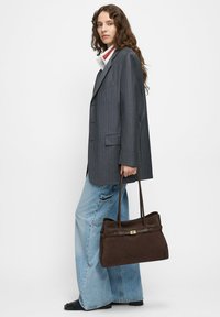 Brązowa zamszowa torebka z podwójnymi uchwytami, prostokątnym kształtem, złotymi akcentami, zestawiona z szarą marynarką w paski i jasnoniebieskimi szerokimi spodniami jeansowymi.