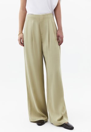 Frau trägt beige, hochtaillierte, weit geschnittene Hosen kombiniert mit schwarzen Schuhen mit quadratischer Spitze und einem weißen Oberteil, steht vor weißem Hintergrund.