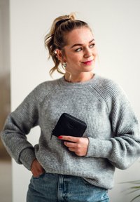 Gray ribbed sweater with raglan sleeves, paired with blue denim jeans. Holding a small black zippered wallet with embossed detailing.
