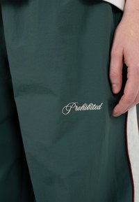 Hånd hvilende ved siden af mørkegrønne bukser med hvid kursiv tekst "Forbudt" nær lårområdet.