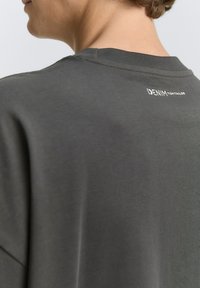 Tricou gri cu o textură moale și netedă, având un guler rotund și un mic logo alb imprimat pe spate.