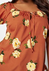 Bluzka w wzór kwiatowy w kolorze spalonym pomarańczowym; posiada marszczony dekolt z wiązaniem oraz duże żółte róże z zielonymi liśćmi.