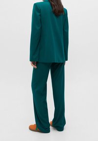 Turkusowy dopasowany garnitur z długimi rękawami i luźnymi spodniami, noszony z pomarańczowymi sneakersami. Gładki, fakturowany materiał, prosty design i swobodny krój.
