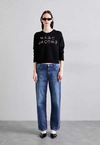 Pulover tricotat negru cu text multicolor "MARC JACOBS", asortat cu blugi drepti albaștri și pantofi negri cu vârf ascuțit.