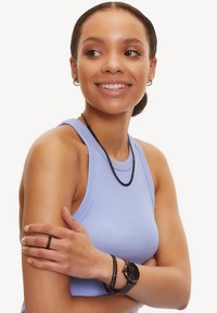 Collana intrecciata nera, bracciale abbinato e anello su una persona che indossa una canotta azzurra chiara. Orologio semplice con quadrante scuro al polso.