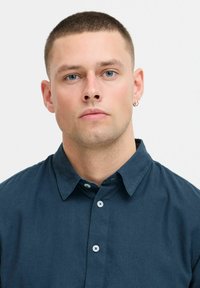 Chemise col bleu foncé en tissu à la texture lisse. Elle présente une fermeture à boutons et des boutons contrastants au niveau du col et sur le devant.