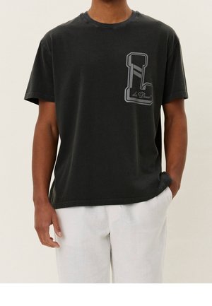 Osoba ubrana w czarną koszulkę z dużym logo "L" i białe spodnie, stojąca z jedną ręką w kieszeni na jednolitym tle.