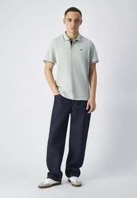 Jasnozielona koszulka polo z czarnym kołnierzykiem i akcentami rękawów, w połączeniu z ciemnoniebieskimi, szerokimi spodniami dżinsowymi i białymi butami. Prosta postawa.