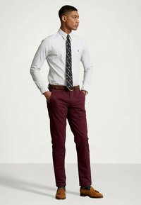 Vīrietis ar svītrotu baltu kreklu, rakstainu tumši zilu kaklasaiti, tumši sarkanām biksēm, brūnu jostu un gaišām frakņu kurpēm, stāv ar rokām kabatās.