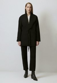 Giacca nera a maniche lunghe con cintura in vita, caratterizzata da un design ispirato al kimono e tasche, abbinata a pantaloni neri a gamba dritta e stivaletti.