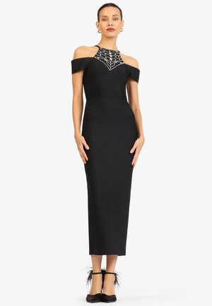 Frau steht in einem figurbetonten schwarzen Midikleid mit Cut-out-Schultern und einem bestickten, hochgeschlossenen Hals, kombiniert mit schwarzen federverzierten Knöchelriemen-Absatzschuhen.