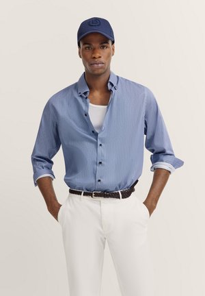 Blaues Langarmhemd mit kleinem Muster, geknöpft, kombiniert mit weißen Hosen und einem geflochtenen schwarzen Gürtel. Marineblaues Barett mit Logodetail.