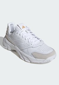 Chaussure de sport blanche avec tige en cuir perforé, accents en suède gris clair et semelle en caoutchouc texturée. Comprend des lacets plats et un col rembourré.