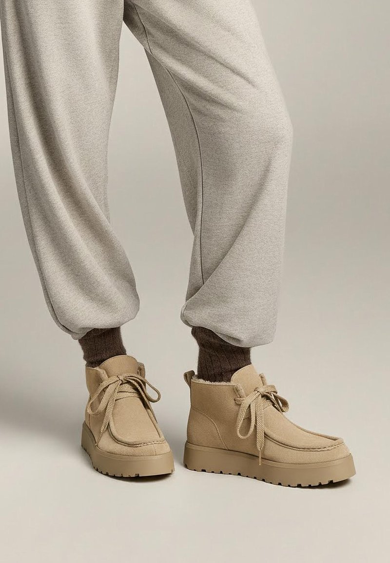 Osoba nosiaca béžové semišové členkové topánky s hrubými podrážkami, viazanými šnúrkami, hnedé rebrované ponožky a svetlošedé voľné tepláky.