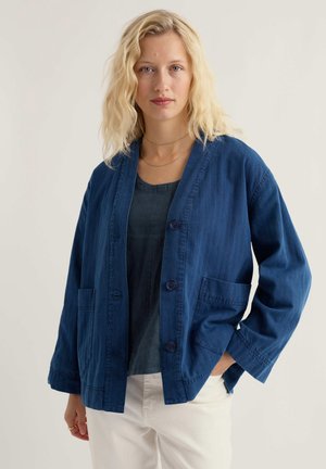 Szőke nő hullámos hajjal, sötétkék, gombos zakót visel fekete felső és fehér nadrág fölött, világos háttér előtt állva.